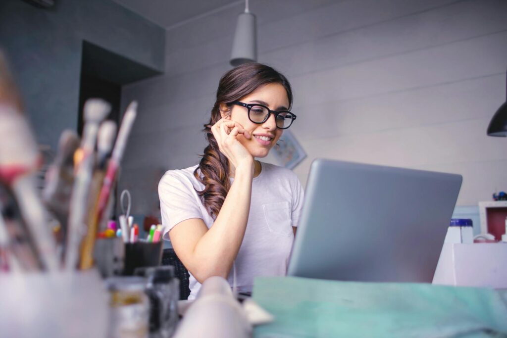 Auxiliar Administrativo e de Recrutamento e Seleção, Remotas A cheerful woman in glasses works remotely on her laptop in a cozy home office setting.