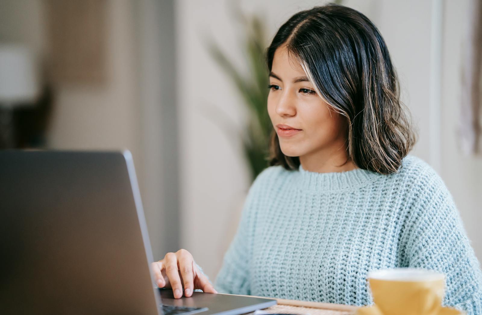 Vagas de Emprego na TIM: 55 Oportunidades Abertas Confident young Latin American woman in casual clothes working remotely on netbook at table near mug with coffee in light apartment with plant with green leaves near wall