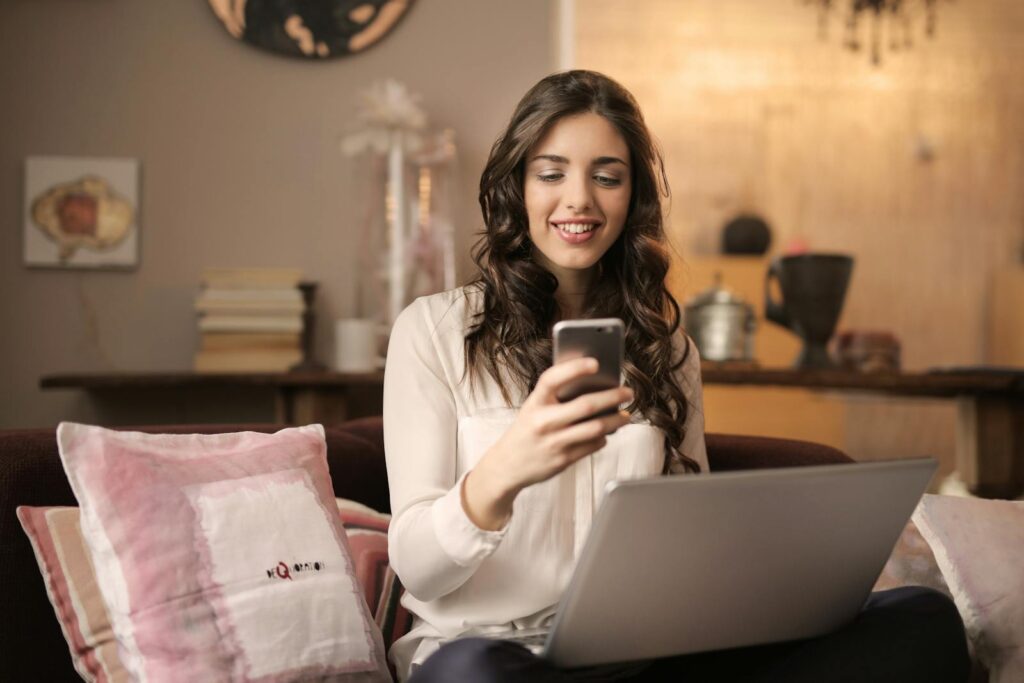 Auxiliar e Assistente em Vagas Híbridas A woman enjoying leisure time using her smartphone and laptop in a cozy living room.